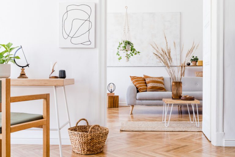 Ett modernt vardagsrum med en grå soffa och randiga kuddar, ett soffbord i trä med torkat gräs, hängande växter, väggkonst och en korg på ett fiskbensmönstrat trägolv. Ett skrivbord och en stol syns i förgrunden.