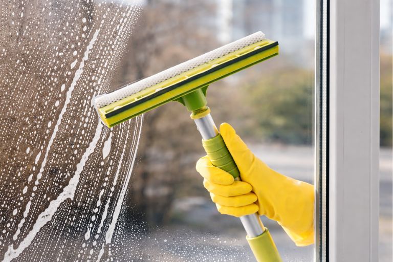 En person med en gul gummihandske använder en grön och svart gummiskrapa för att rengöra ett fönster.